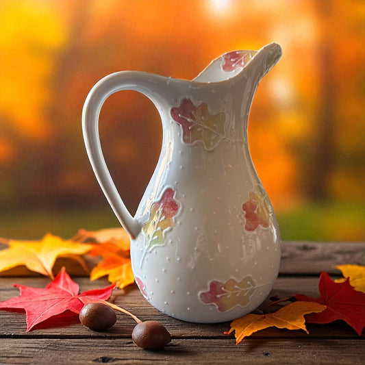 White ceramic elegant pitcher with leaf patterns on a wooden surface with autumn leaves and background.