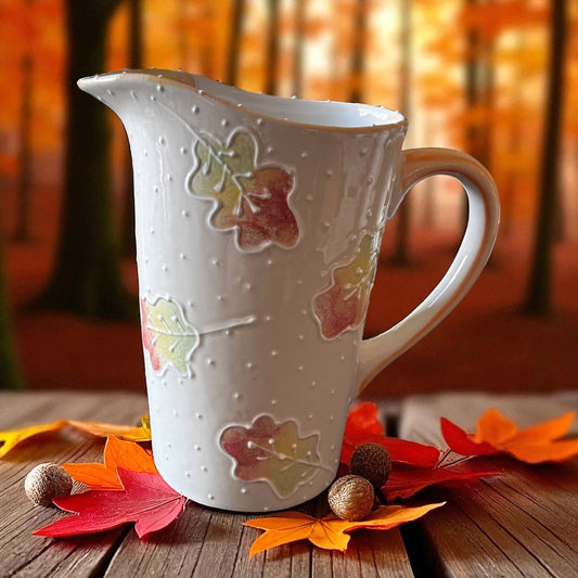 White ceramic milk pitcher with autumn leaf design on a wooden surface with fall leaves and nuts.