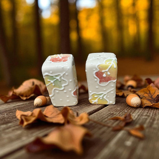 Two painted salt and pepper shakers with autumn-themed designs on a wooden surface with leaves and acorns.