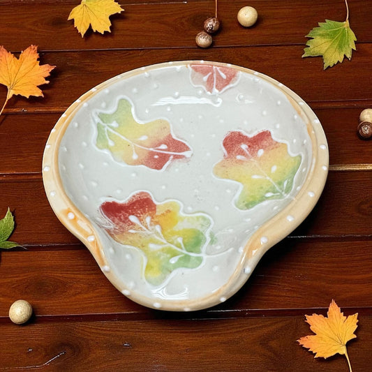Decorative ceramic spoon rest with autumn leaf design on a wooden surface with fall leaves and acorns.