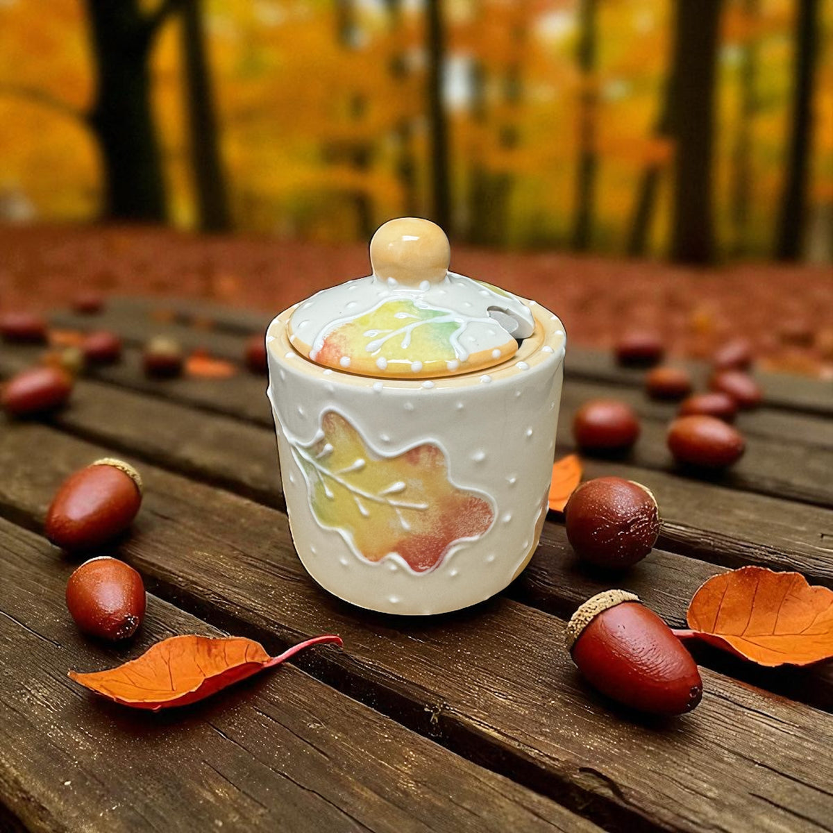Decorative sugar jar with lid featuring autumn leaves on a wooden surface with fall foliage in the background. 