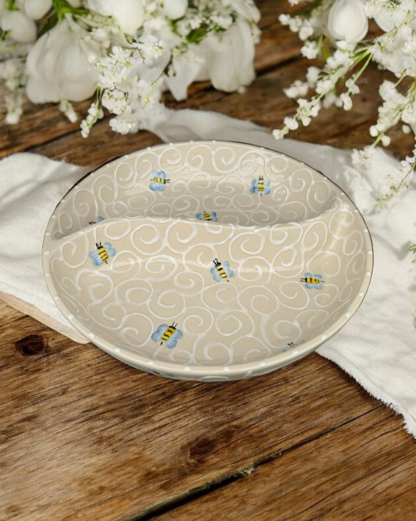 A ceramic two-compartment serving Bowl with a white base decorated with a cute bee pattern, set on a white wooden surface with potted flowers in the background.