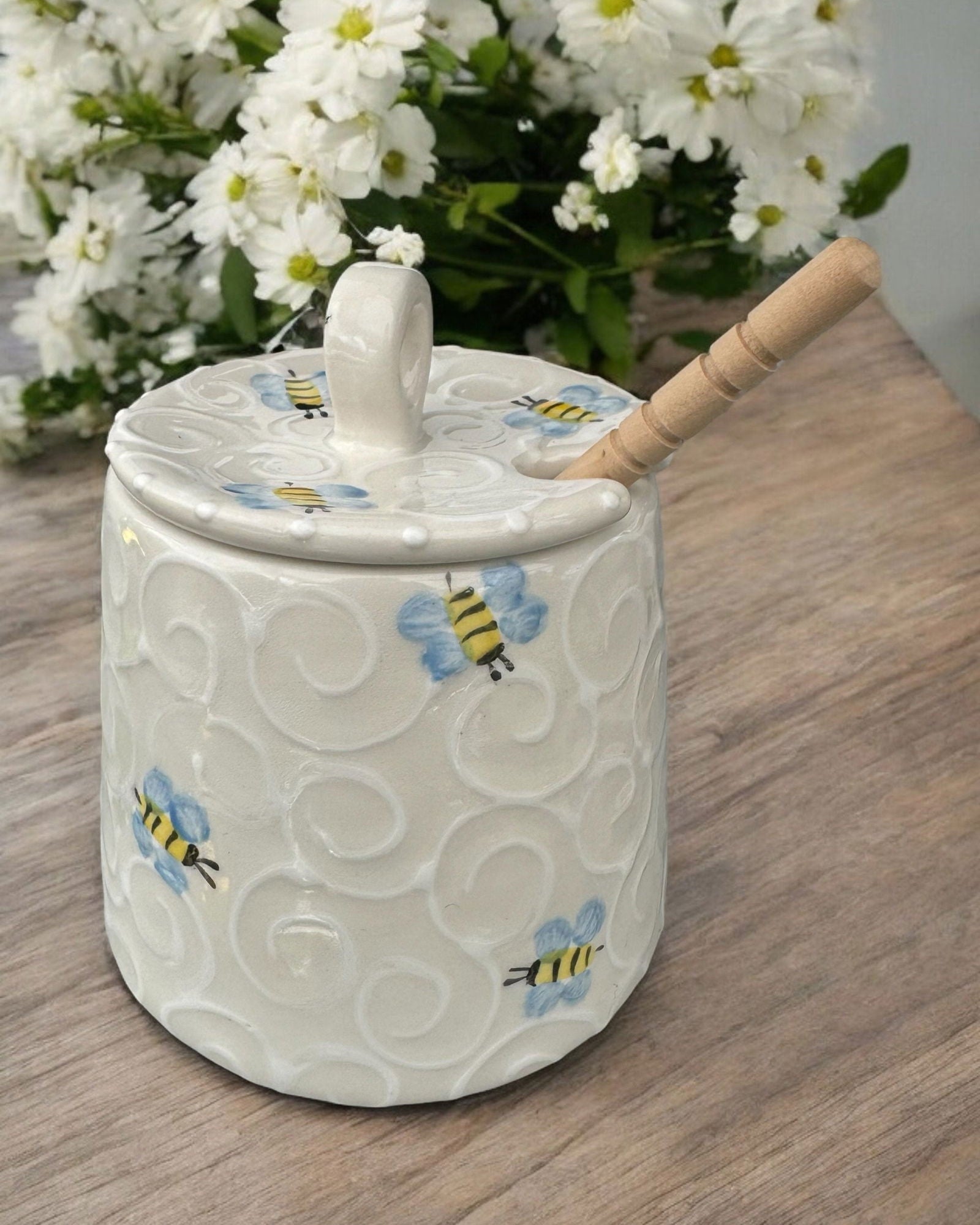 A cream-colored  ceramic honey pot jar with a lid and a wooden spoon, decorated with colorful wildflower and insect motifs, set on a rustic wooden table.