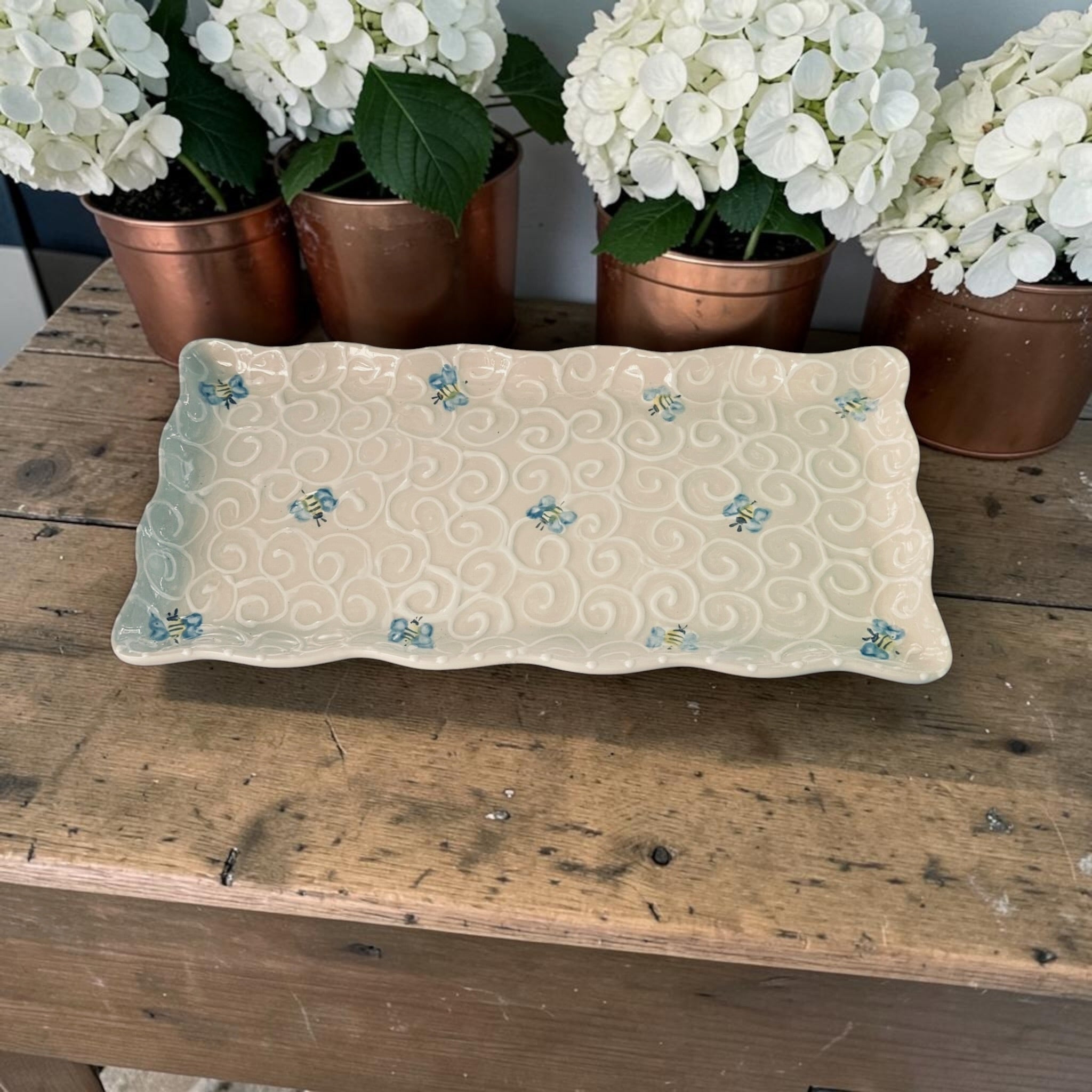Decorative tray with bee patterns on a wooden surface with potted white flowers in the background. Ladybird Ceramics Canada