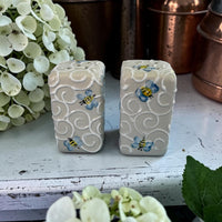 Two ceramic salt and pepper with bee designs on a wooden surface with flowers and pots in the background.