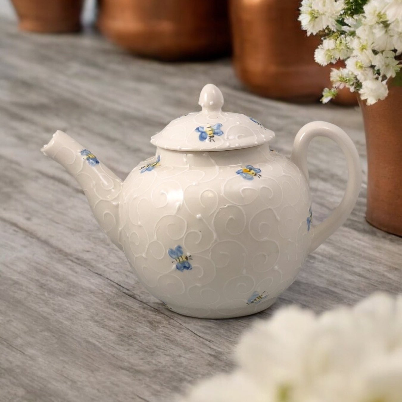 A white ceramic teapot with hand-painted bee designs, displayed on a wooden surface with a bouquet of white flowers beside it.