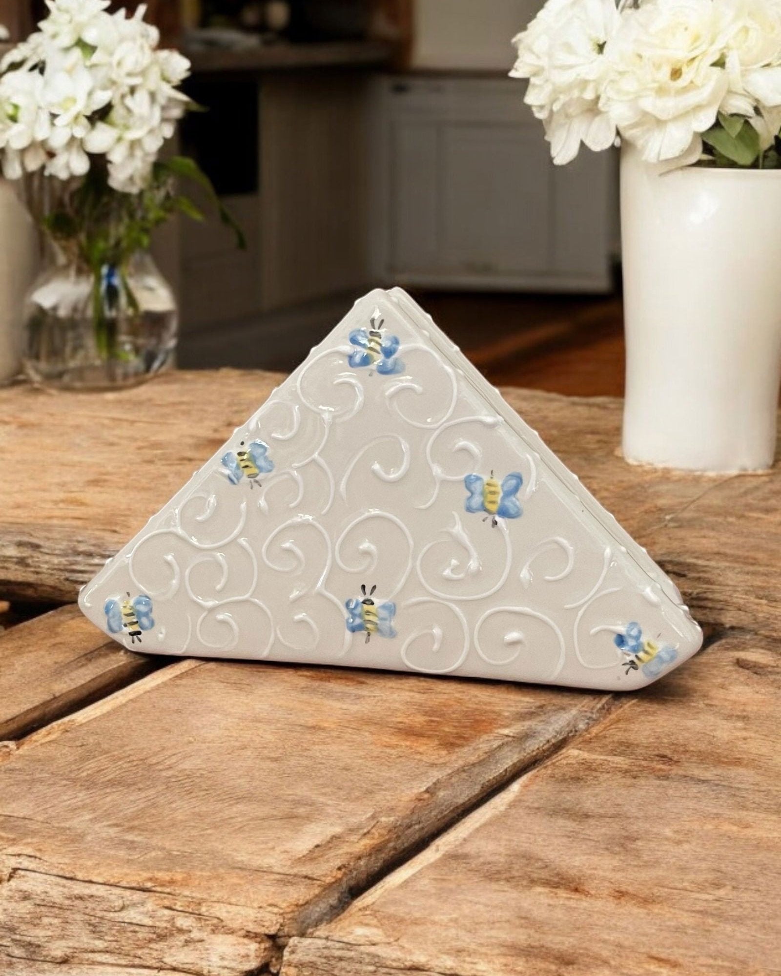 A triangular white ceramic napkin holder with painted bee and floral patterns displayed on a wooden table.