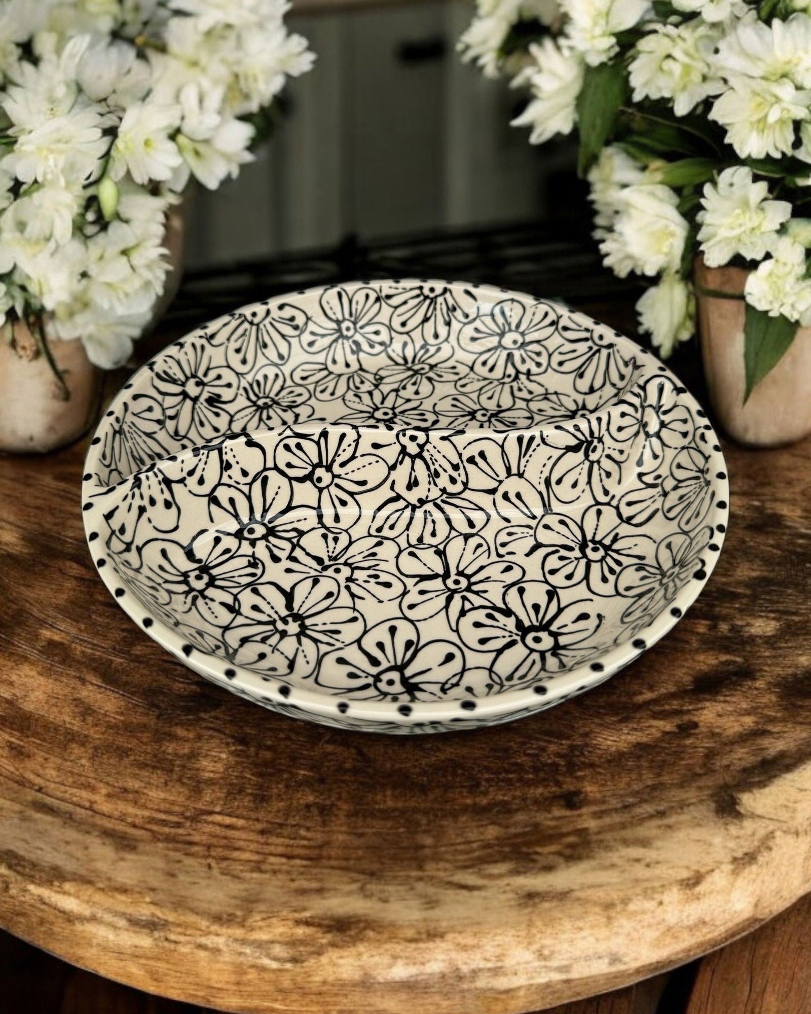 A ceramic two-compartment serving Bowl with black and white floral pattern, set on a white wooden surface with potted flowers in the background.