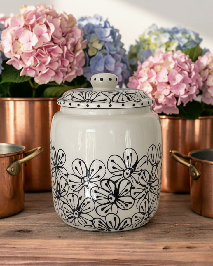 A white ceramic canister with a black floral pattern and matching lid, displayed on a wooden table with copper pots and pink, blue, and green hydrangeas in the background.