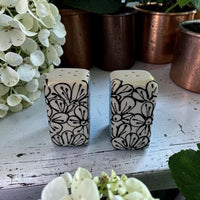 Two ceramic salt and pepper shakers with floral patterns on a wooden surface with plants in the background.