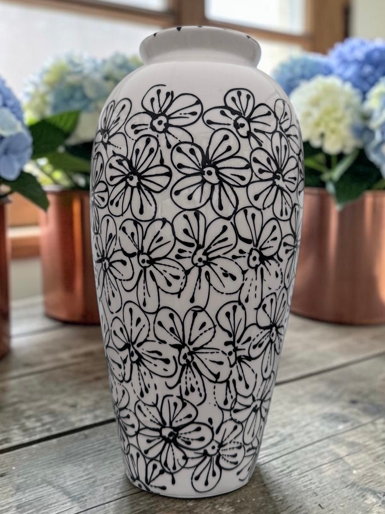 A white ceramic vase with a black hand-drawn floral pattern, standing on a rustic wooden table with blurred potted flowers in the background.