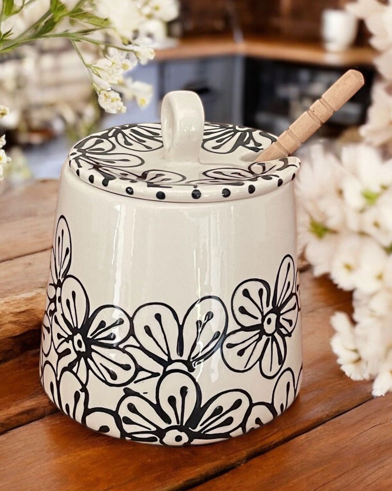 A white ceramic honey pot with black and white floral designs, a wooden spoon, and white flowers on a wooden table.