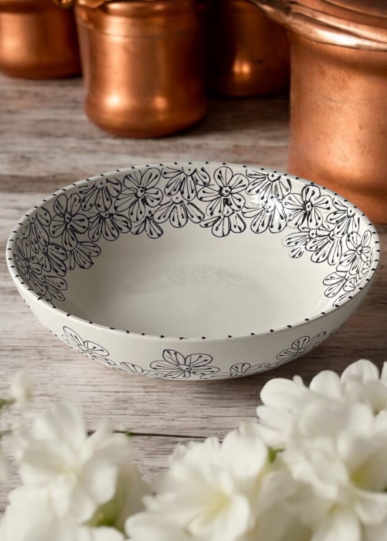 A hand-painted ceramic serving bowl with floral patterns, displayed on a wooden table with daisies in the foreground and copper cookware in the background.