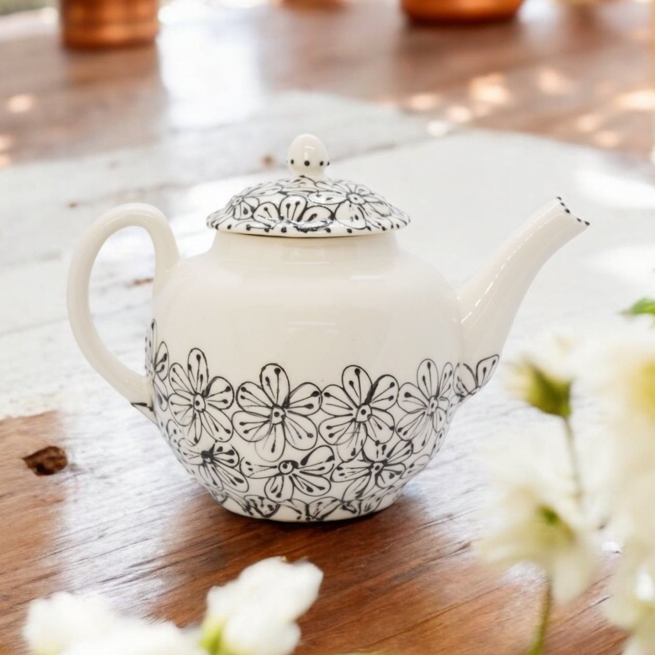 A black and white ceramic teapot with hand-painted floral designs, displayed on a wooden surface with a bouquet of white flowers beside it.