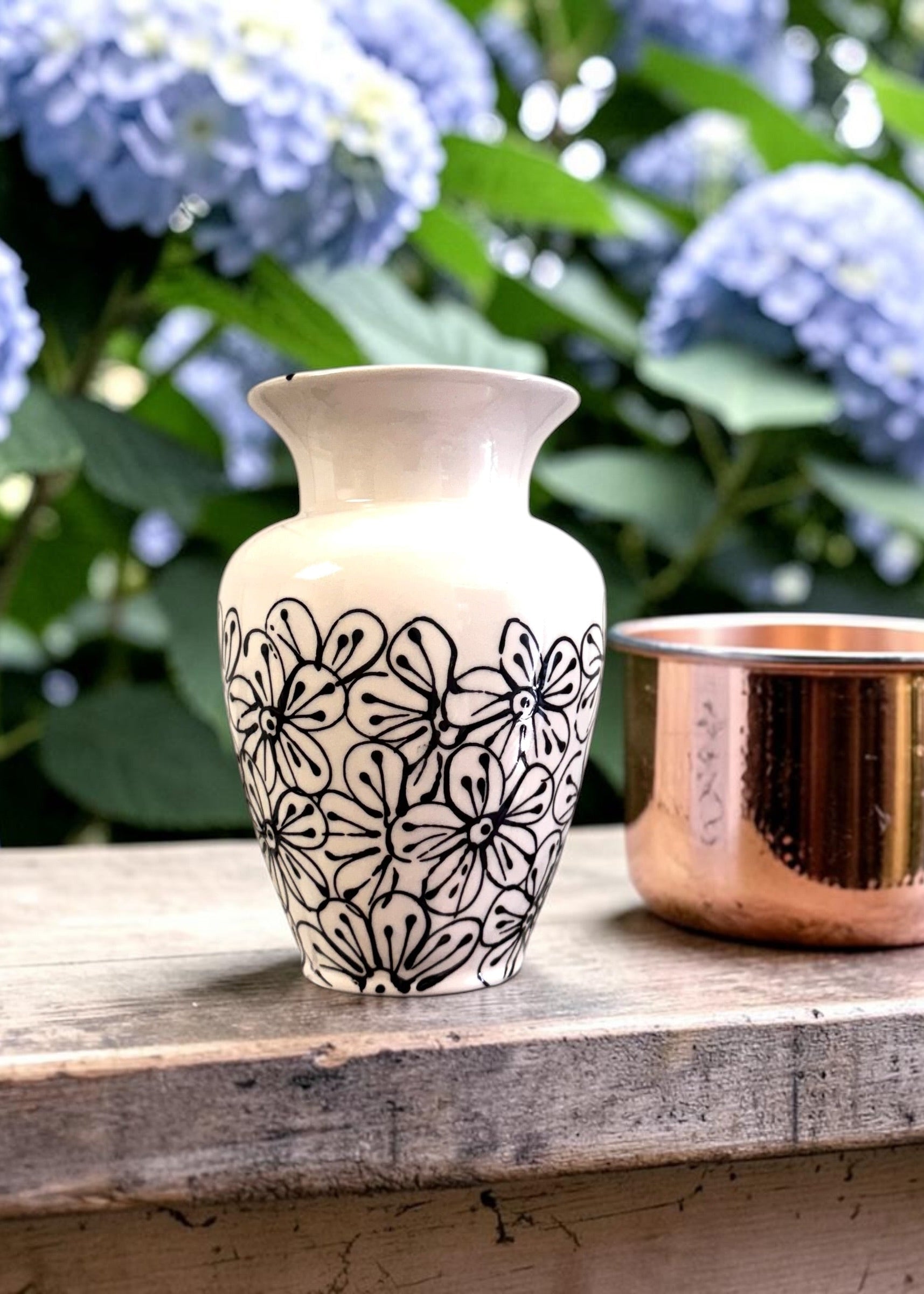 White vase with black floral patterns on a wooden surface with blue flowers in the background
