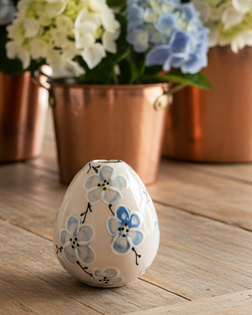 A small ceramic egg-shaped vase with blue and white floral patterns sits on a wooden table, with blurred copper flower pots and hydrangeas in the background.