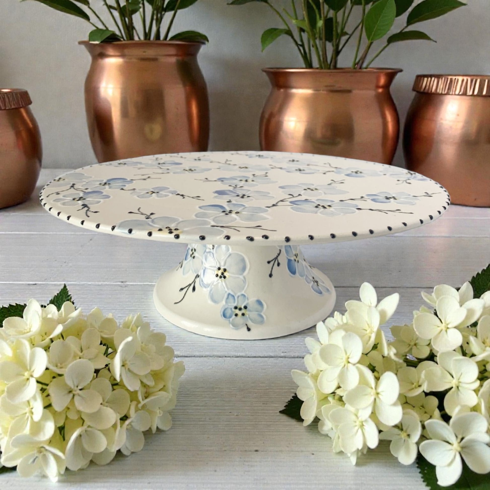 Decorative cake stand with blue cherry blossom floral design on a white surface with copper planters and white flowers.