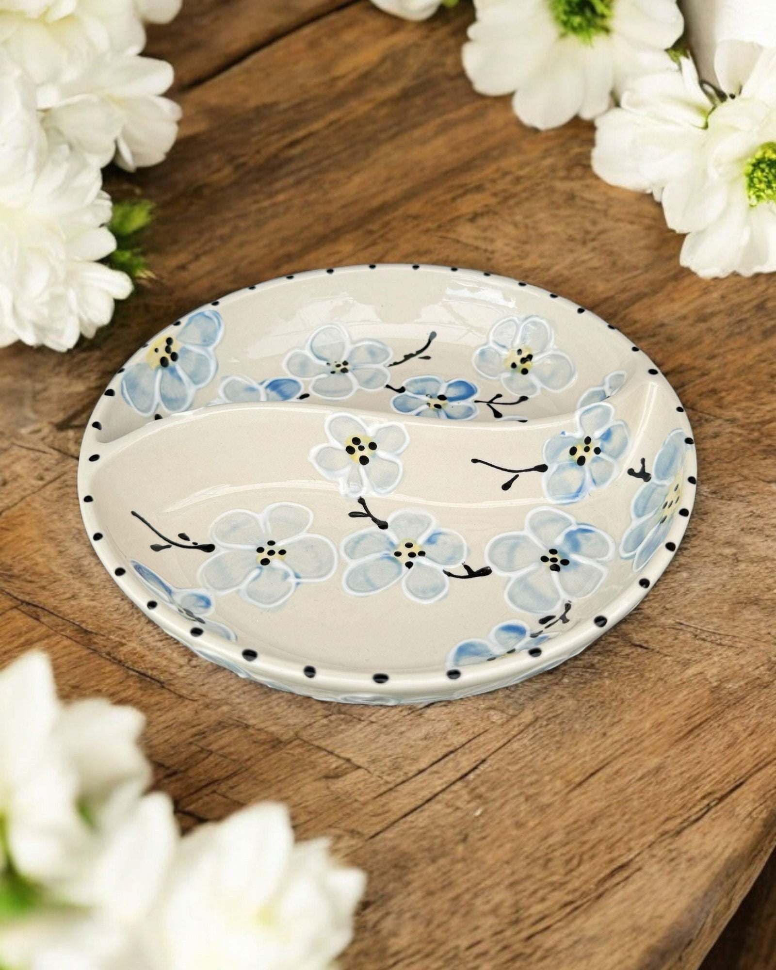A ceramic two-compartment serving Bowl with a white base decorated with a colorful blue floral pattern, set on a white wooden surface with potted flowers in the background.