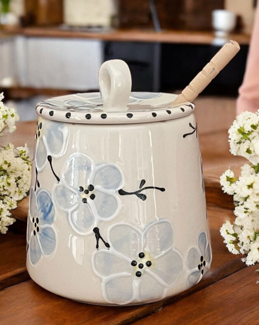 A ceramic honey pot with a lid, a small handle, and a wooden spoon resting inside. The pot is decorated with light blue and white flowers and black dots, set on a wooden table with white flowers on either side.