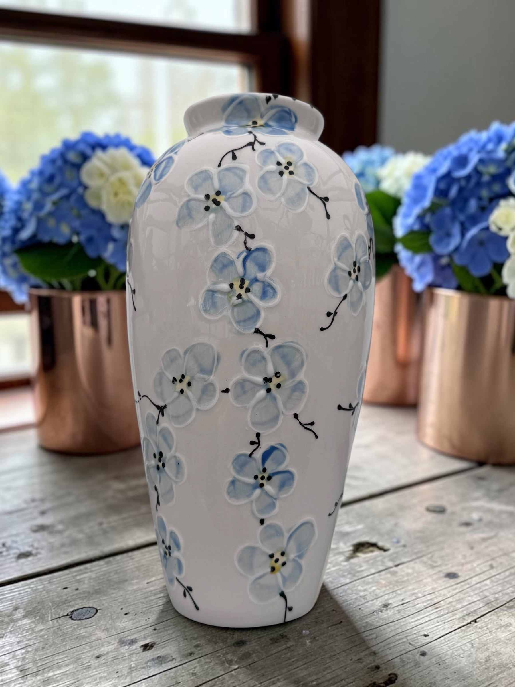 A white ceramic vase decorated with hand-painted blue forget-me-not flowers and black stems, displayed on a rustic wooden table with copper flower pots in the background.