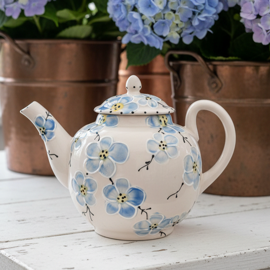 A cream-colored ceramic teapot decorated with a blue and yellow floral pattern, set on a white wooden table in front of copper planters filled with blue hydrangeas.