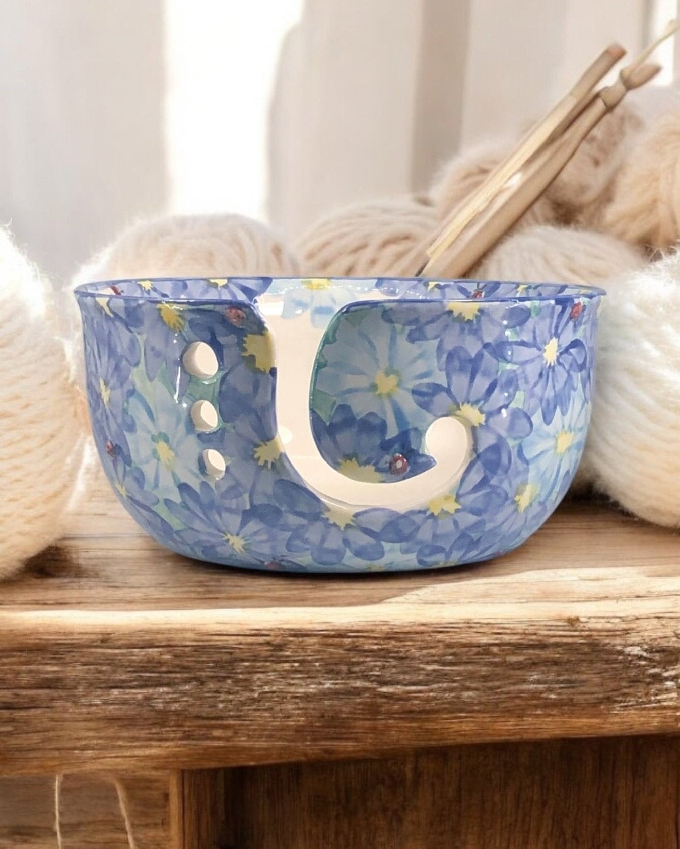 A hand-painted ceramic knitting bowl with blue daisy floral designs, containing yarn balls and knitting tools, placed on a wooden surface. Ladybird Ceramics Canada