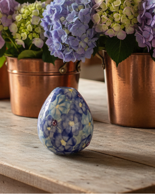 A small decorative ceramic egg-shaped vase with a blue and white floral pattern sits on a rustic wooden table, with copper planters and purple hydrangeas blurred in the background.