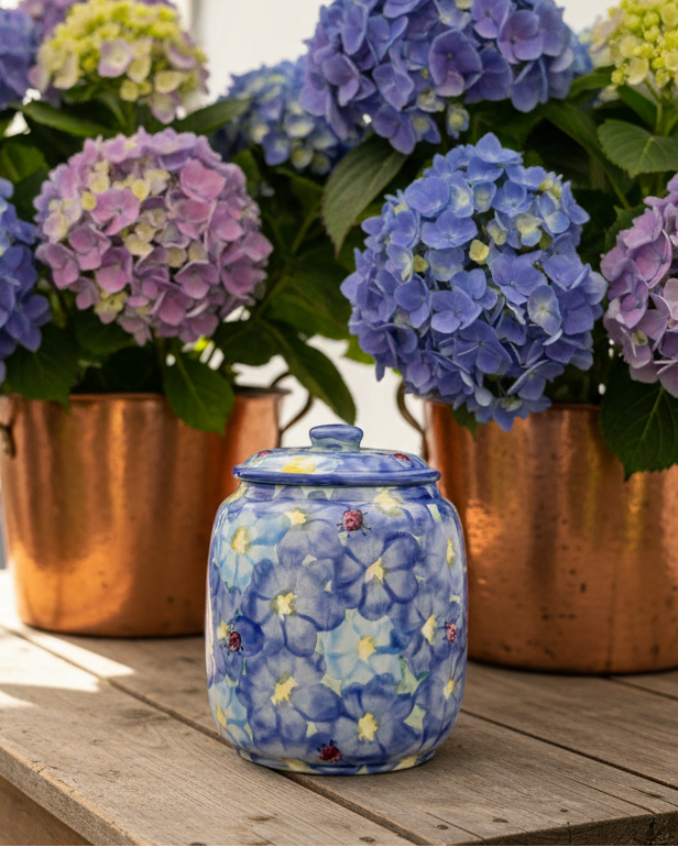 A small blue ceramic jar with a matching lid, decorated with a floral pattern and ladybugs, sits on a wooden table in front of potted blue and purple hydrangeas in copper planters.
