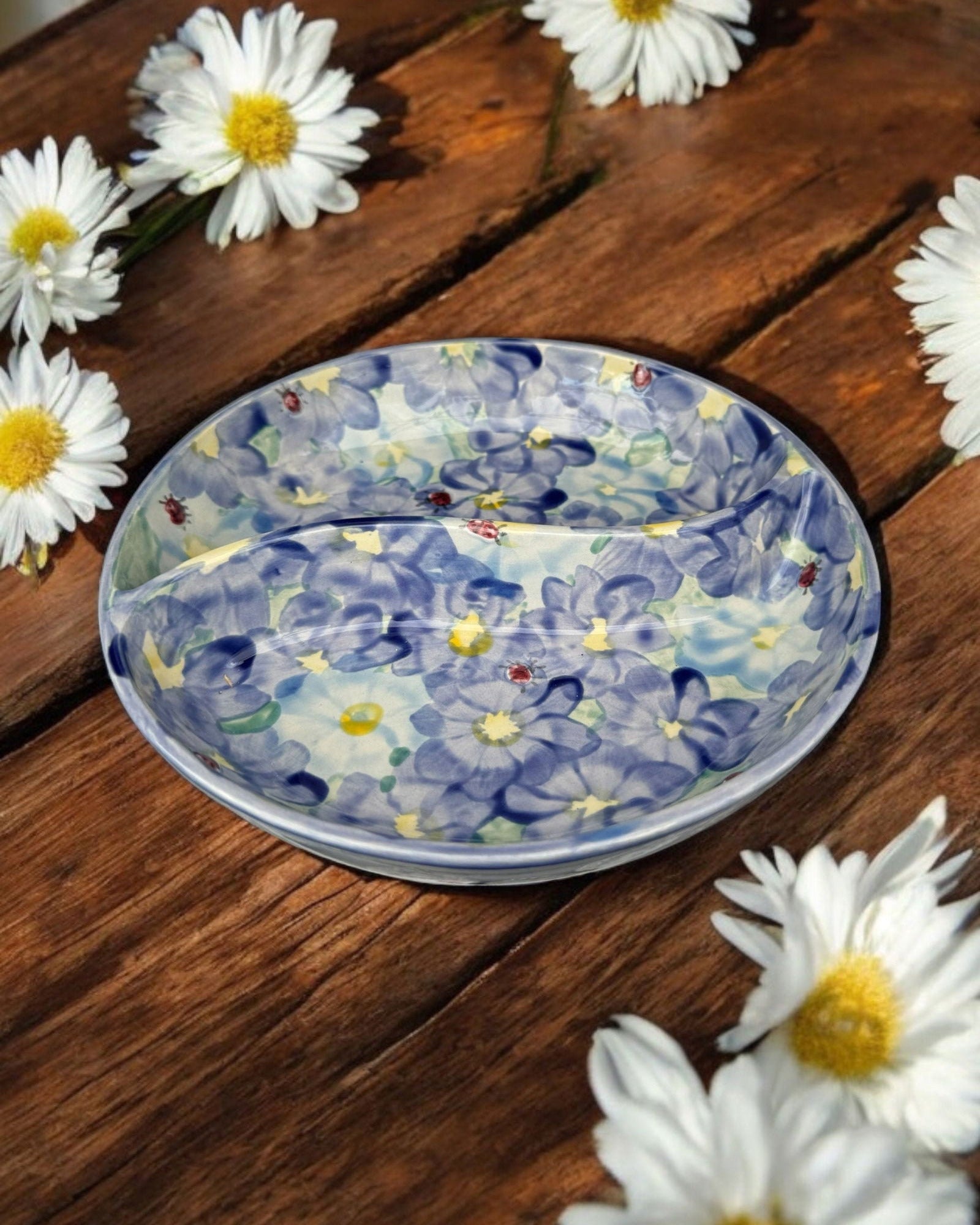 A ceramic two-compartment serving Bowl with a white base decorated with a colorful blue floral pattern, set on a white wooden surface with potted flowers in the background.