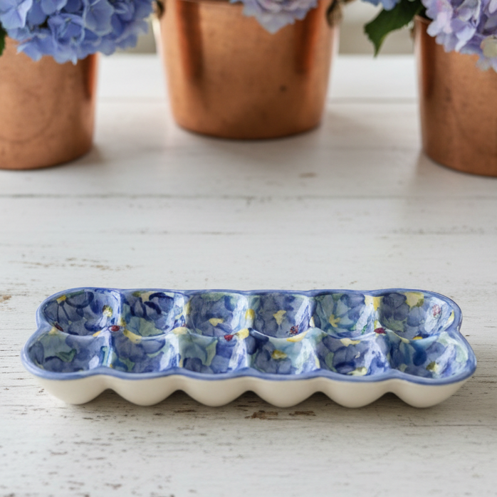 A ceramic egg-shaped dish with a blue floral pattern and white base, placed on a white wooden table with blurred blue hydrangeas in copper pots in the background.