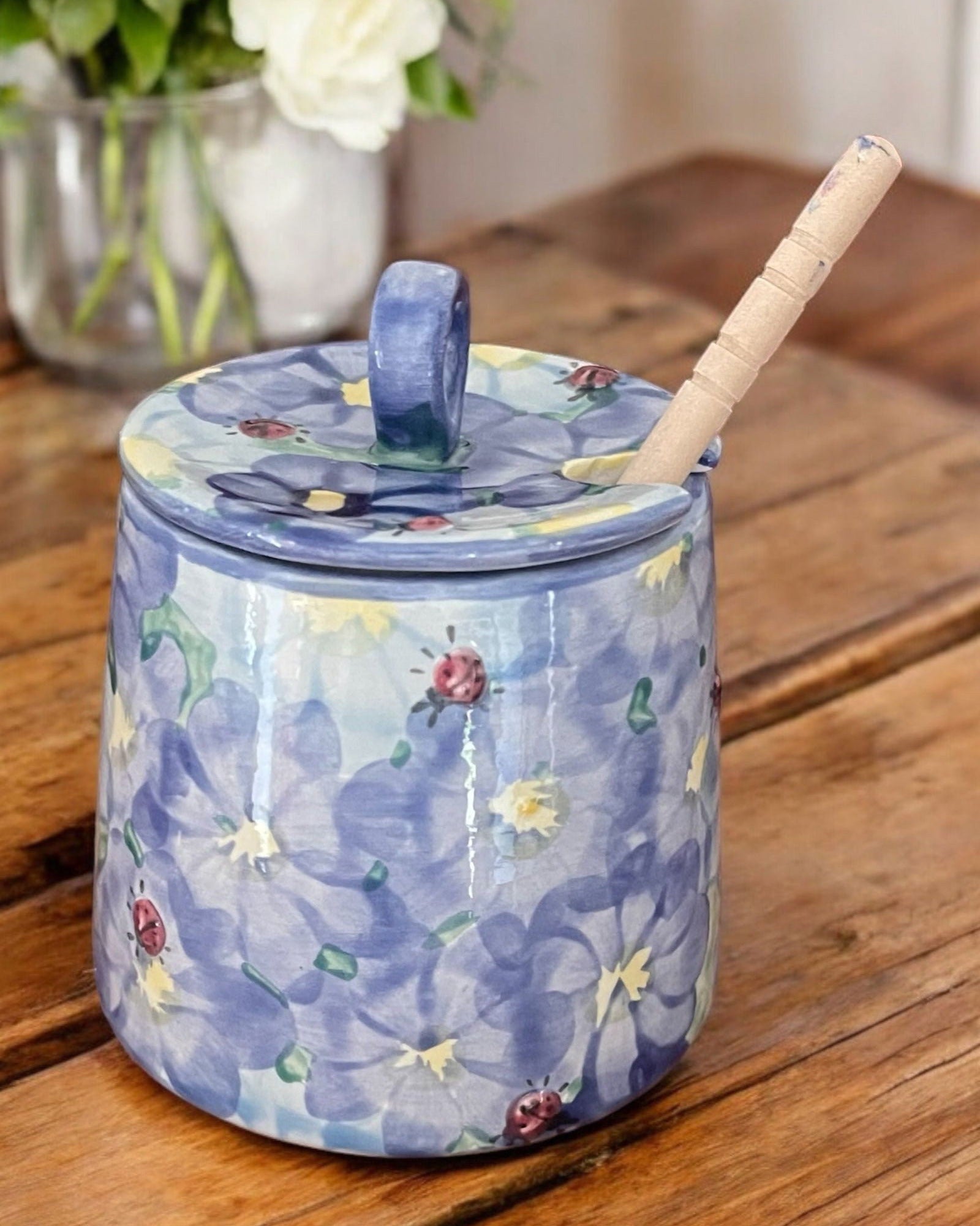 A cream-colored  ceramic honey pot jar with a lid and a wooden spoon, decorated with colorful wildflower motifs, set on a rustic wooden table. 