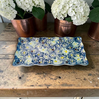 Decorative rectangular plate with floral pattern on a wooden surface with potted plants in the background.