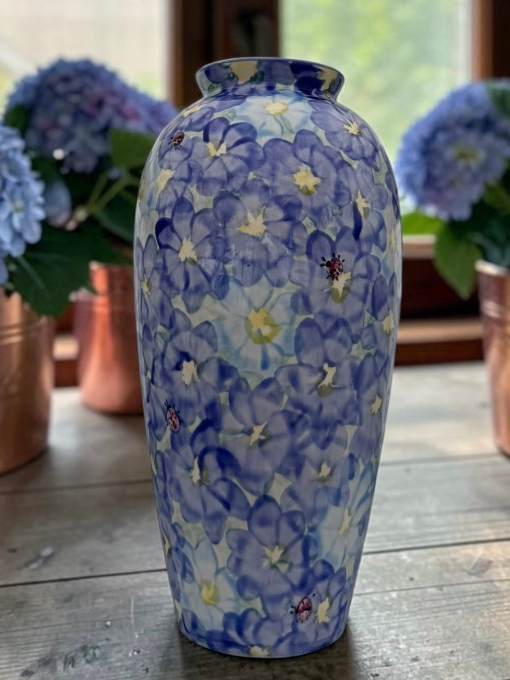 A ceramic vase with a blue and white floral pattern, featuring purple-blue and white flowers with small yellow centers and tiny ladybug details, displayed on a rustic wooden table with blurred hydrangea bouquets in the background.