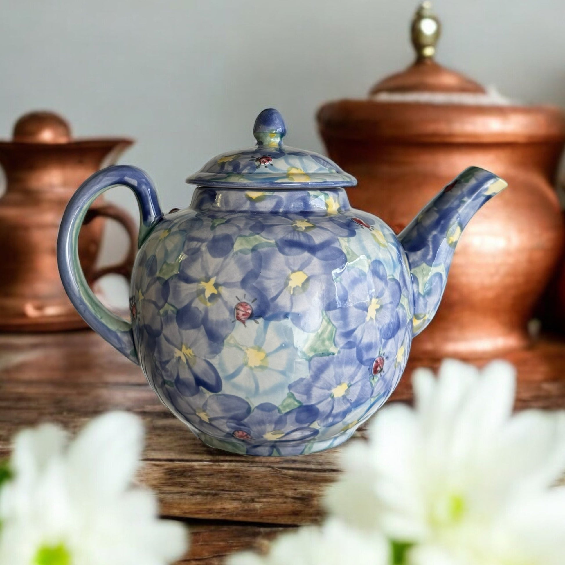 A white ceramic teapot with hand-painted floral designs, displayed on a wooden surface with a bouquet of white flowers beside it.