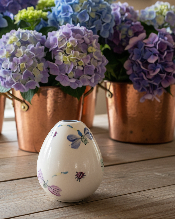 A small white ceramic egg-shaped vase with hand-painted ladybugs and flowers sits on a wooden table in front of copper buckets filled with blue and purple hydrangeas.