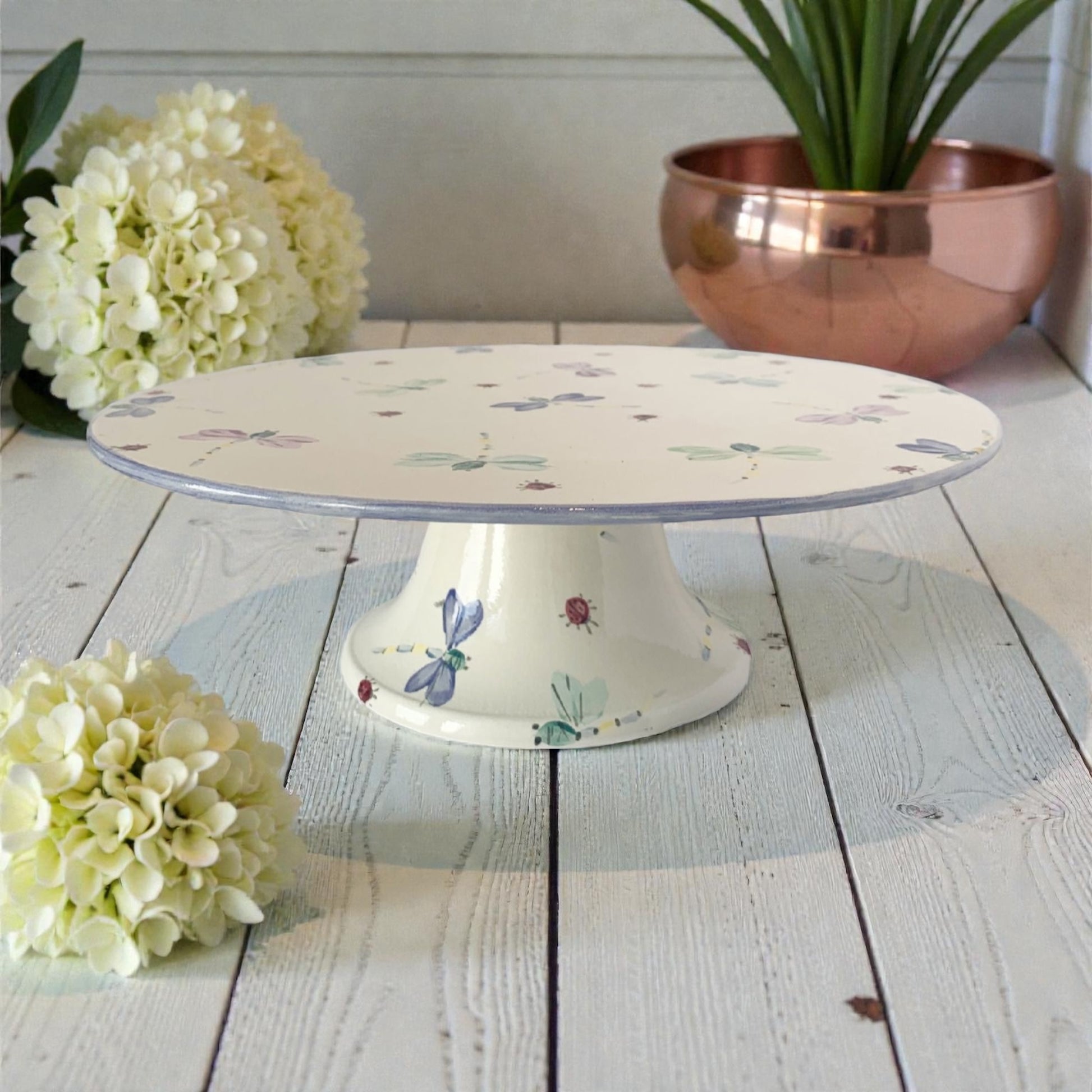 Decorative cake stand with dragonfly and ladybugs  on a wooden surface with flowers and a copper bowl in the background.