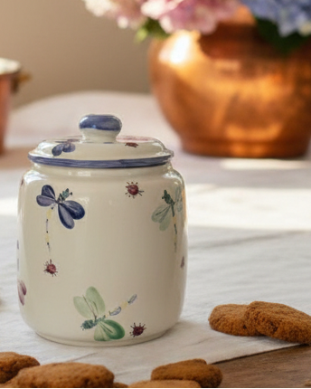 A ceramic canister with a lid, decorated with painted dragonflies and small ladybugs on a cream-colored glaze, set on a table with cookies in the foreground and a blurred flower arrangement in the background.