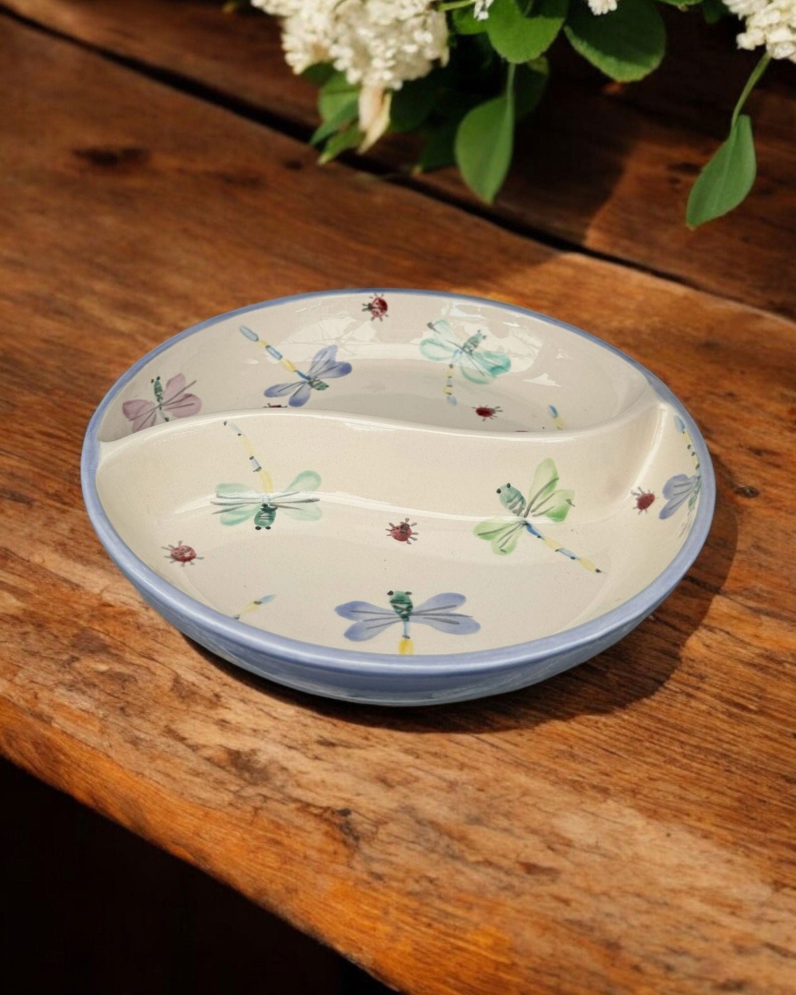 A ceramic two-compartment serving Bowl with a white base decorated with a colorful floral and dragonfly pattern, set on a white wooden surface with potted flowers in the background.
