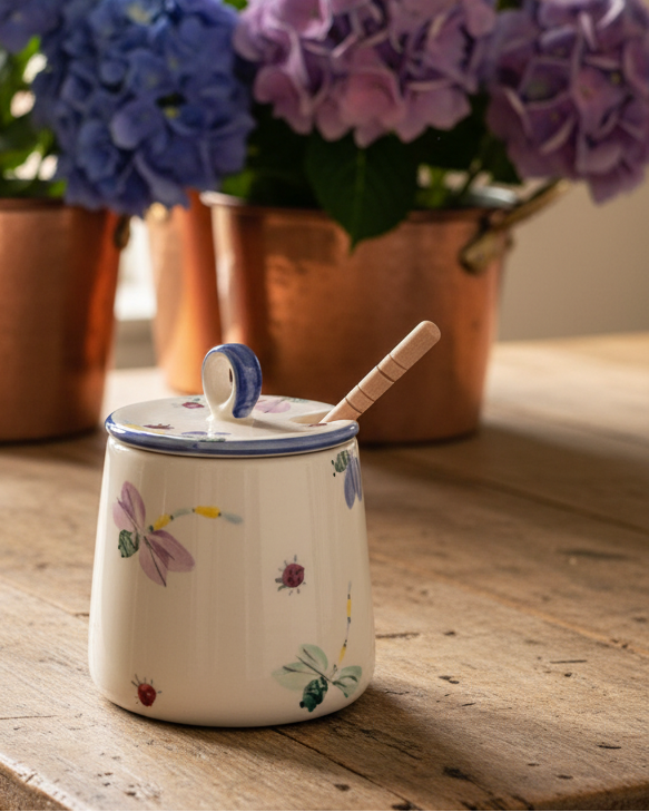 A cream-colored  ceramic honey pot jar with a lid and a wooden spoon, decorated with colorful wildflower and insect motifs, set on a rustic wooden table with blurred purple and blue hydrangeas in the background.