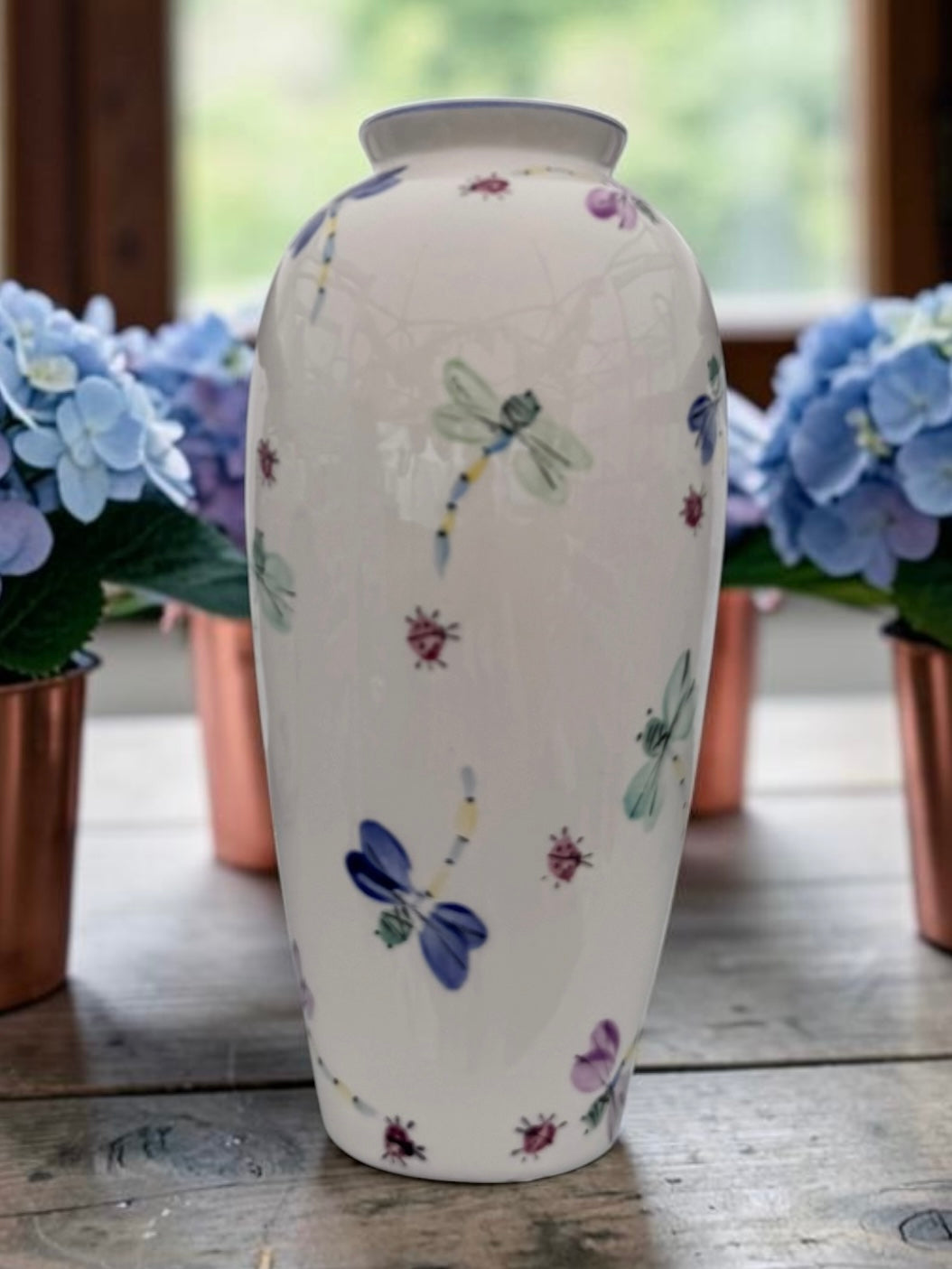 A white ceramic vase decorated with hand-painted dragonflies and small flowers, displayed on a rustic wooden table with blue hydrangeas in the background.