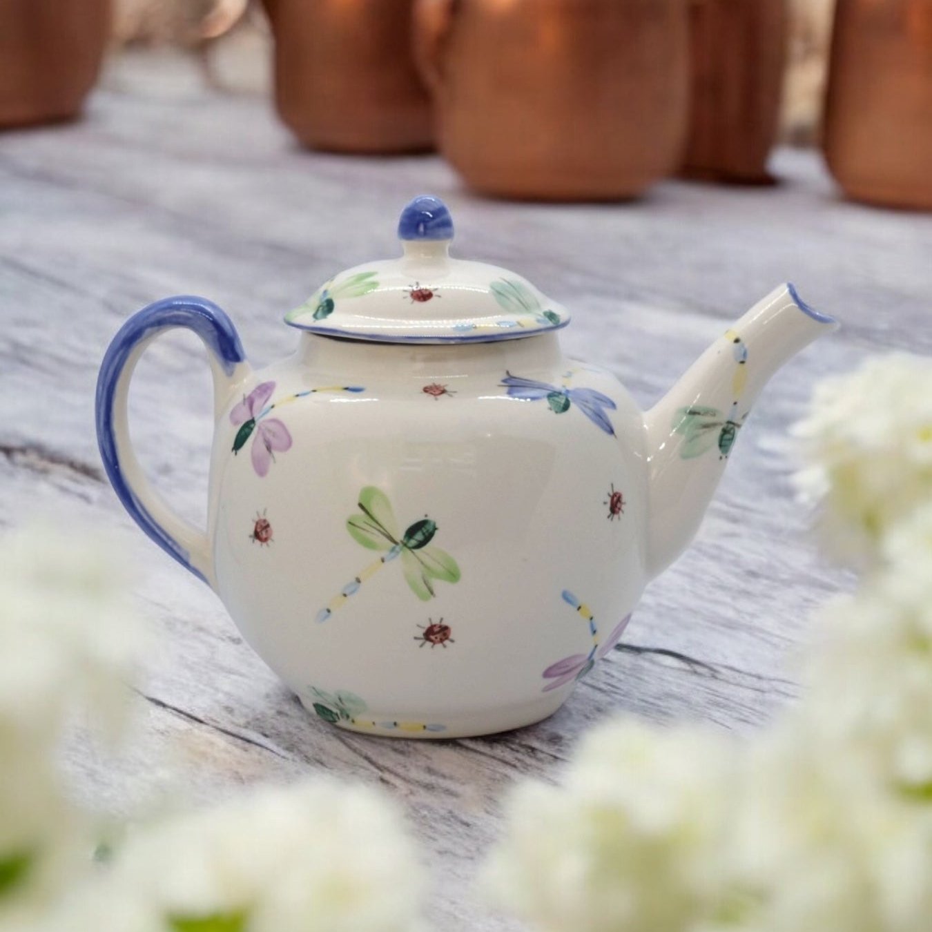 A white ceramic teapot with hand-painted floral and dragonfly designs, displayed on a wooden surface with a bouquet of white flowers beside it.