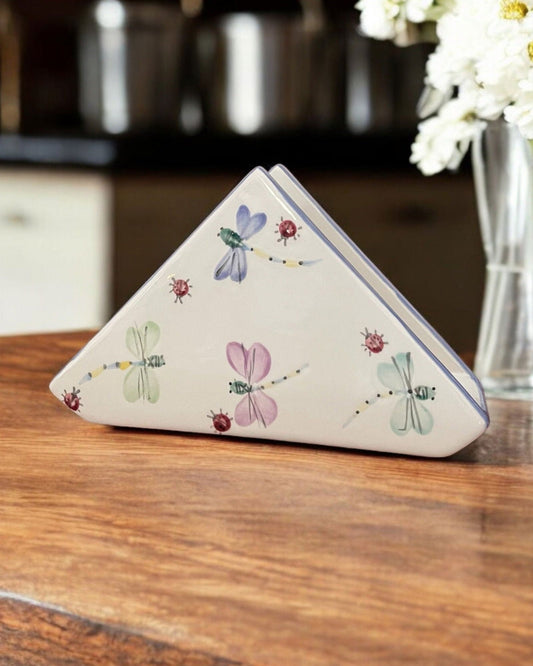 A triangular white ceramic napkin holder with embossed floral patterns and small colorful dragonfly designs, displayed on a wooden table.