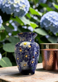 Decorative vase with floral pattern on a wooden surface with copper cups and hydrangeas in the background