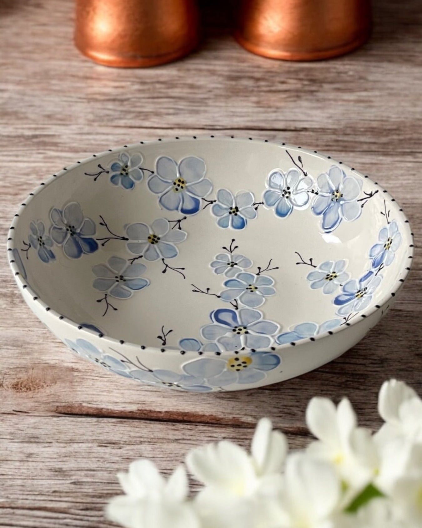 A hand-painted ceramic serving bowl with floral patterns, displayed on a wooden table with daisies in the foreground and copper cookware in the background.