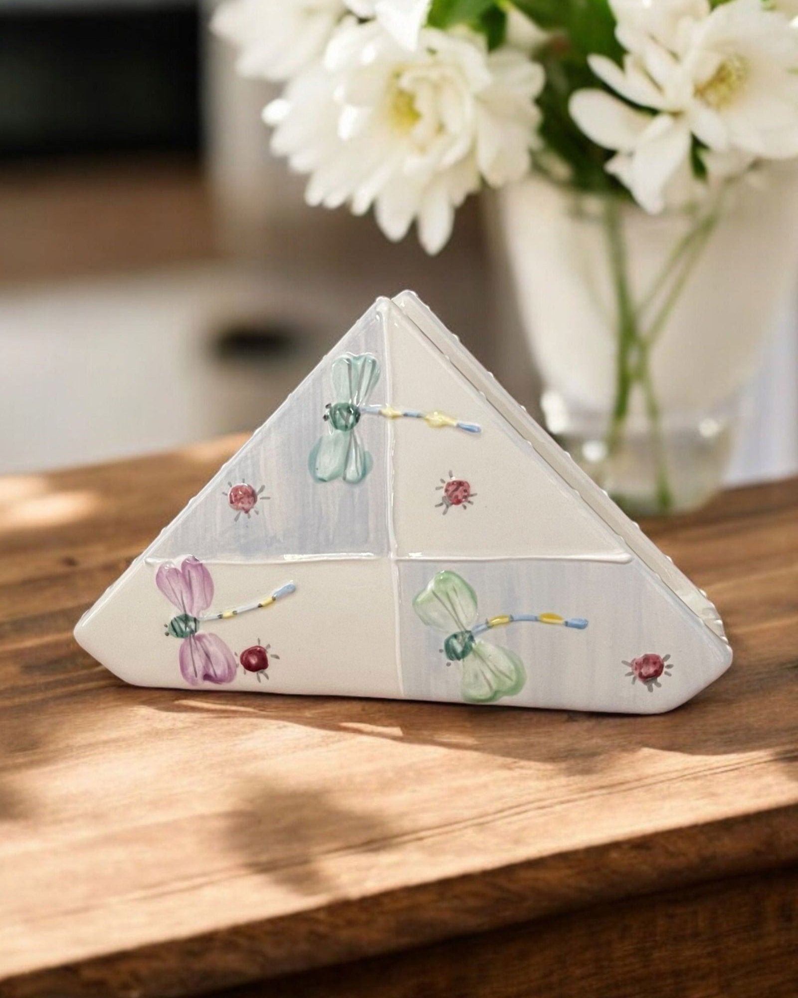A triangular white ceramic napkin holder with painted floral and dragonfly patterns displayed on a wooden table.