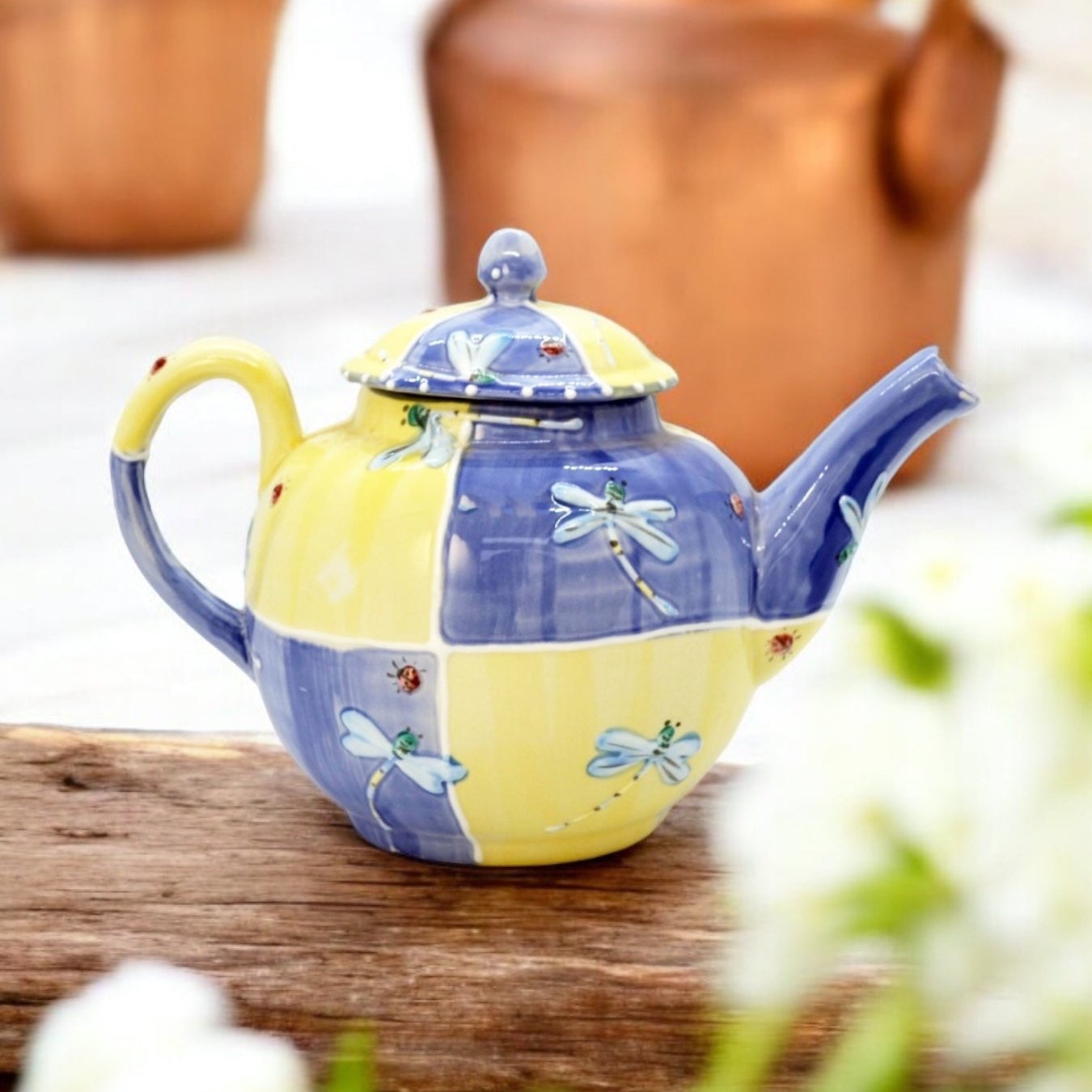 A blue and yellow ceramic teapot with hand-painted floral designs, displayed on a wooden surface with a bouquet of white flowers beside it.