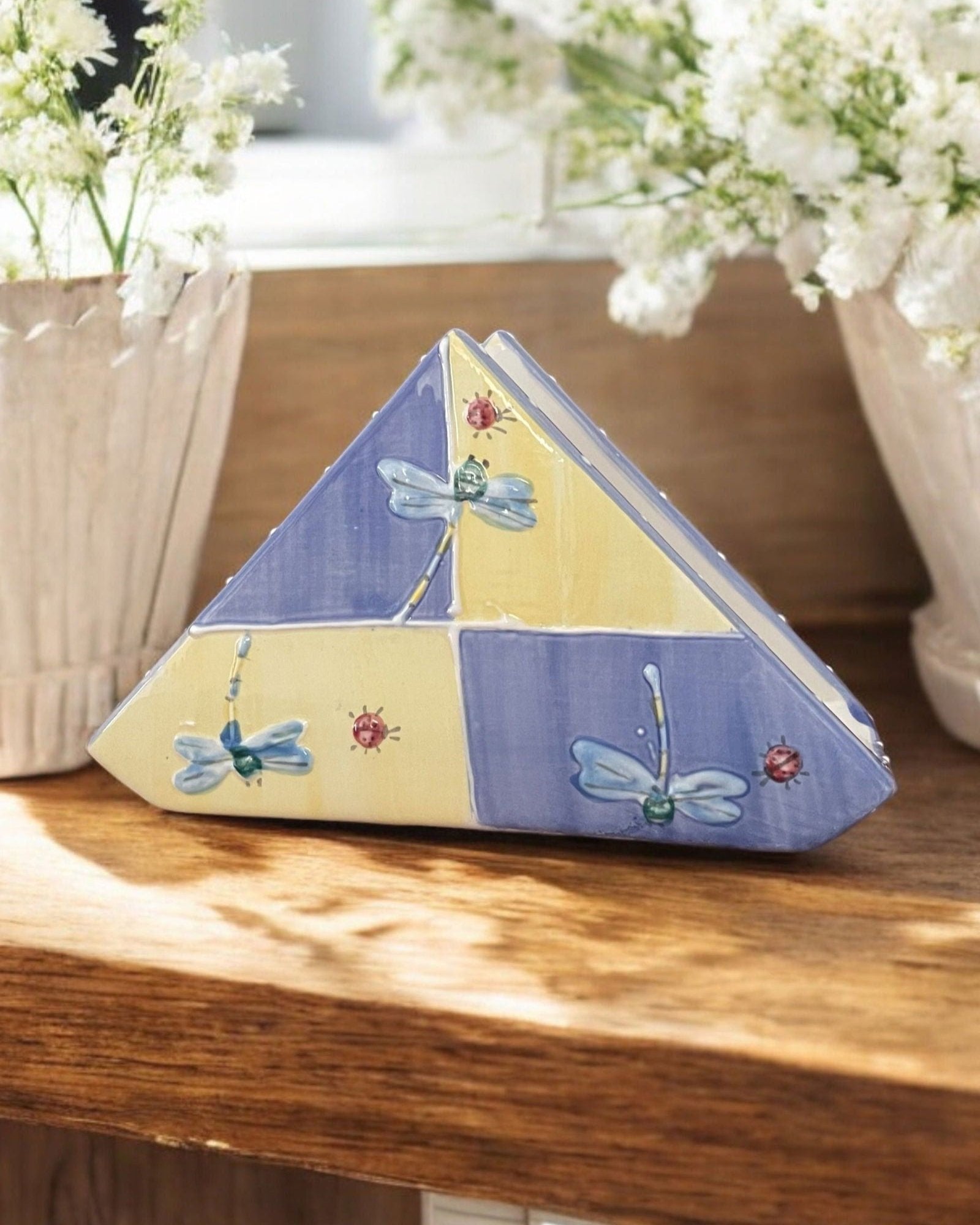 A triangular white ceramic napkin holder with painted floral and dragonfly patterns displayed on a wooden table.