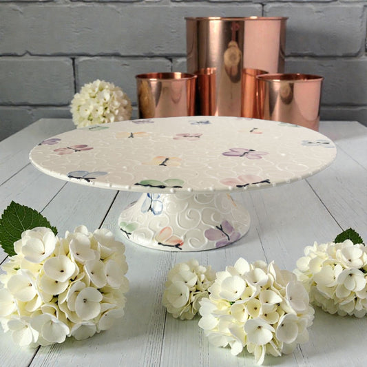 Decorative cake stand with painted butterfly design on a white surface with flowers and copper containers in the background.