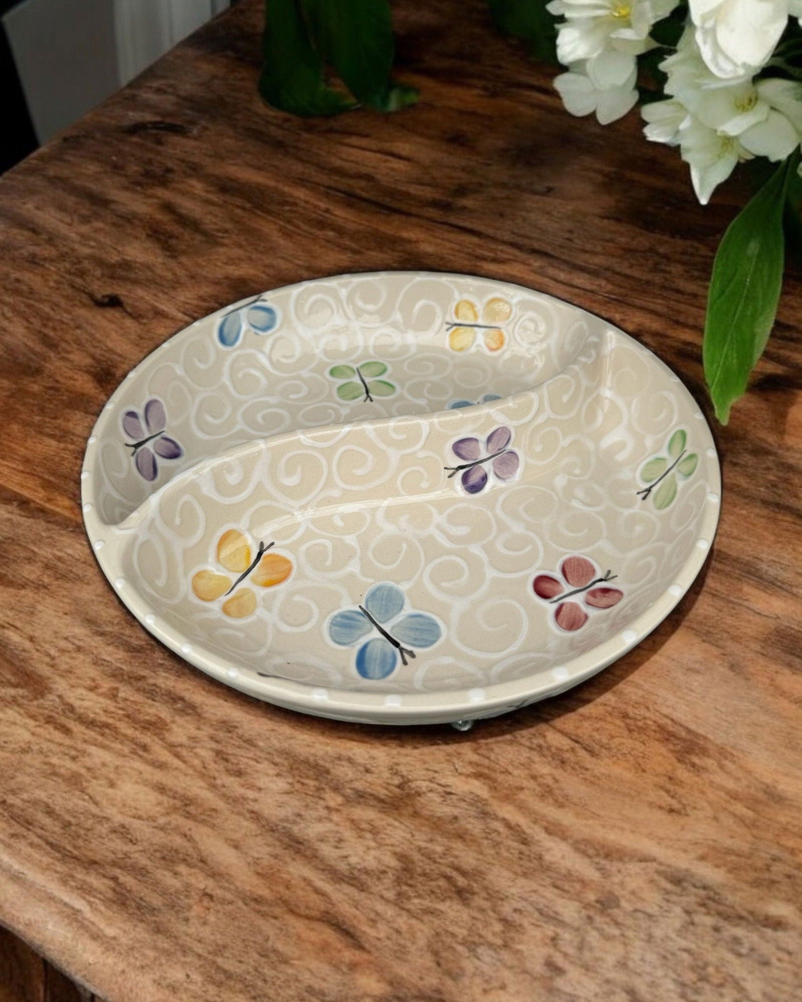 A ceramic two-compartment serving Bowl with a white base decorated with a colorful butterfly pattern, set on a white wooden surface with potted flowers in the background.