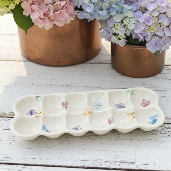A white ceramic egg-shaped tray with butterfly and floral motifs, displayed on a white wooden table with copper pots of pink, blue, and purple hydrangeas in the background.